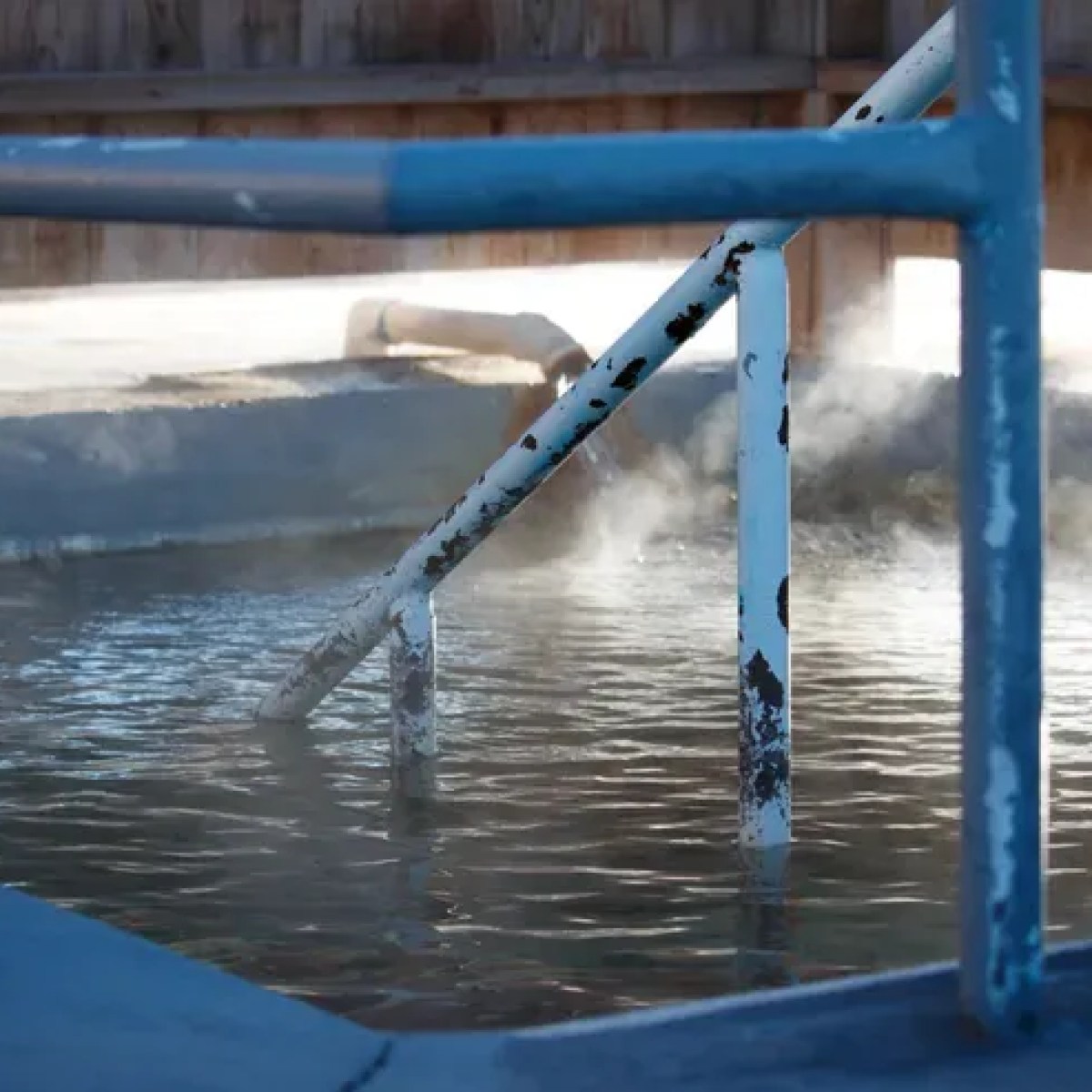 Outdoor hot spring with metal railing and steam rising from water.
