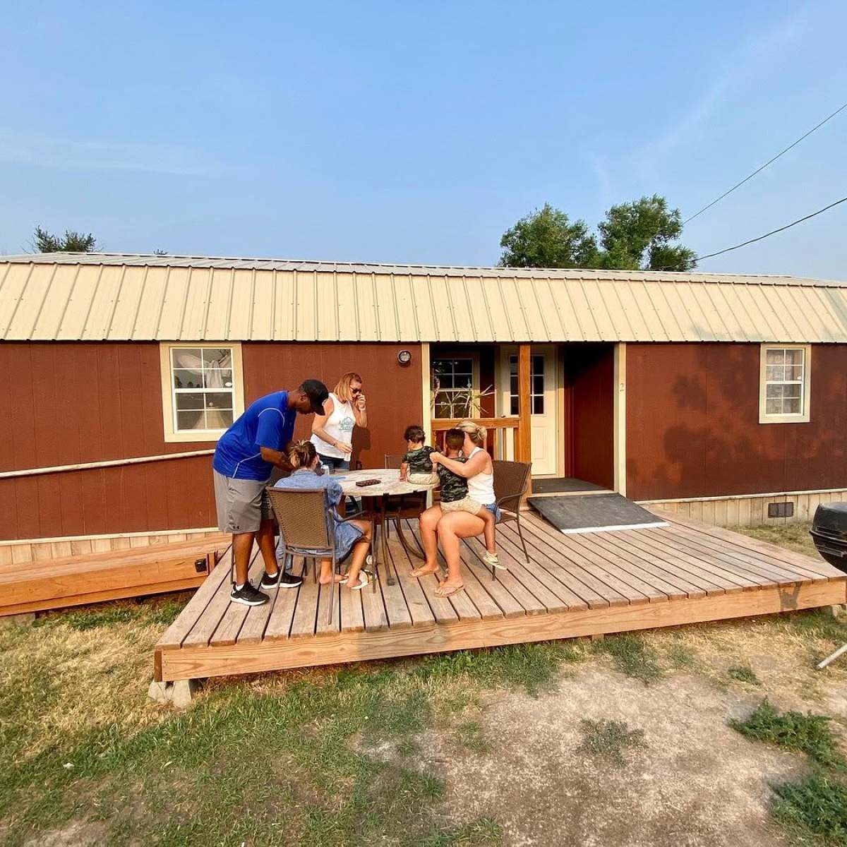 Group of people sitting and standing on wooden deck in front of brown cabin.
