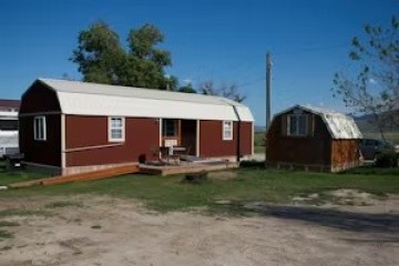 Small red cabin with a porch next to a matching shed on green grass.