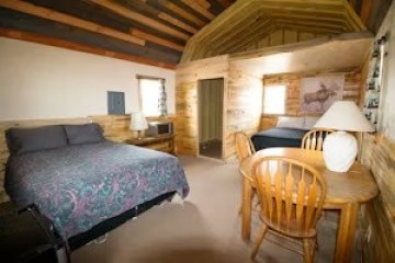 Cozy cabin room with two beds, wooden walls, a small table, chair, and a lamp.