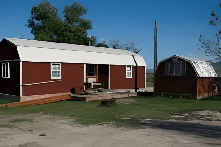 Red cabin and smaller shed on grassy area, with trees and blue sky in background.