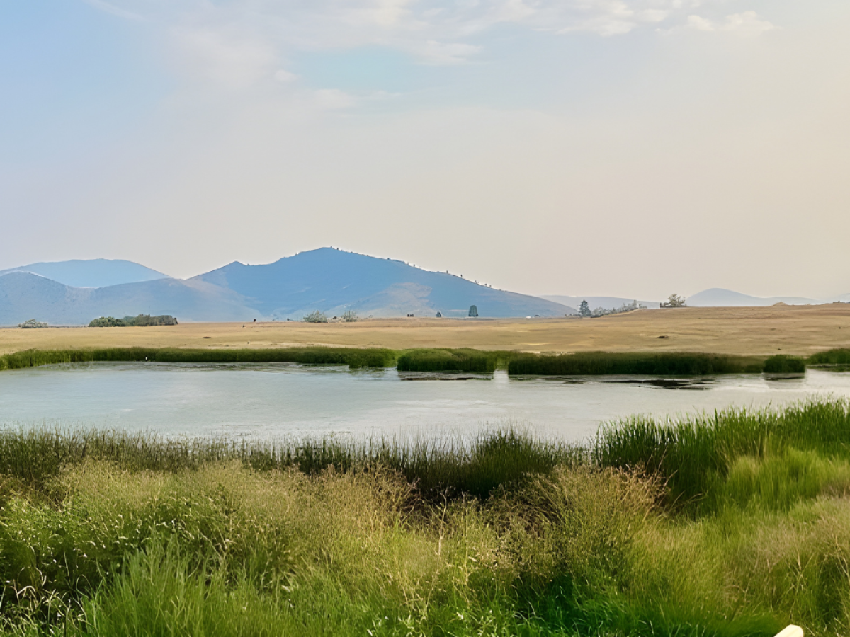 A round outdoor bathtub overlooking a pond and mountains under a blue sky.