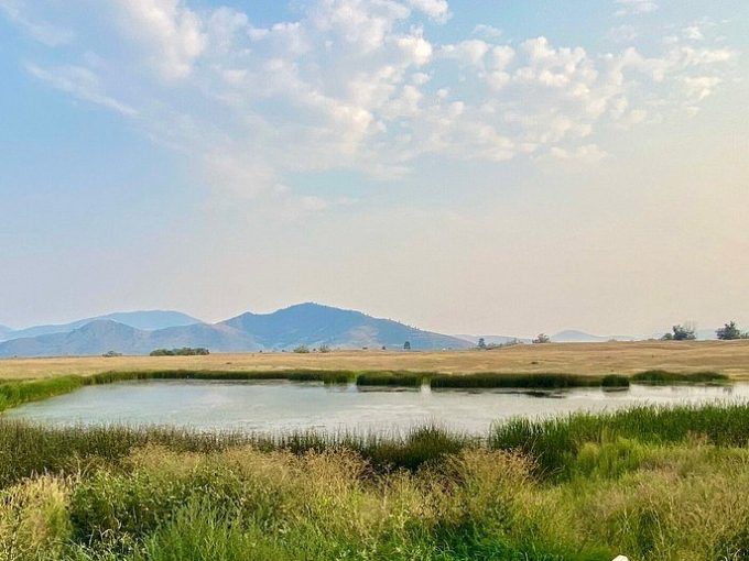 Small pool with bench overlooks grassy field and distant hills under a cloudy sky.