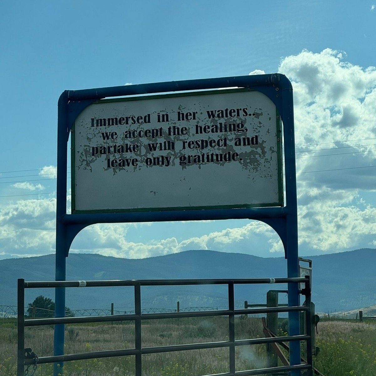 Sign by a rural road with mountains in the background under a blue sky.