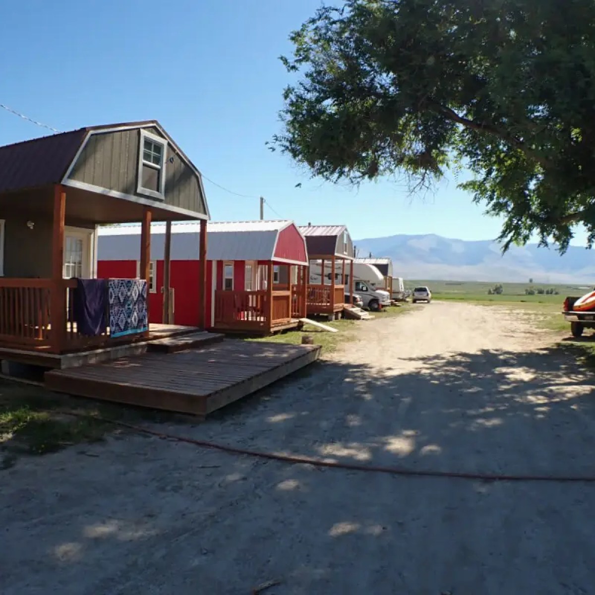 Small cabins and trailers under a tree with mountains in the background.