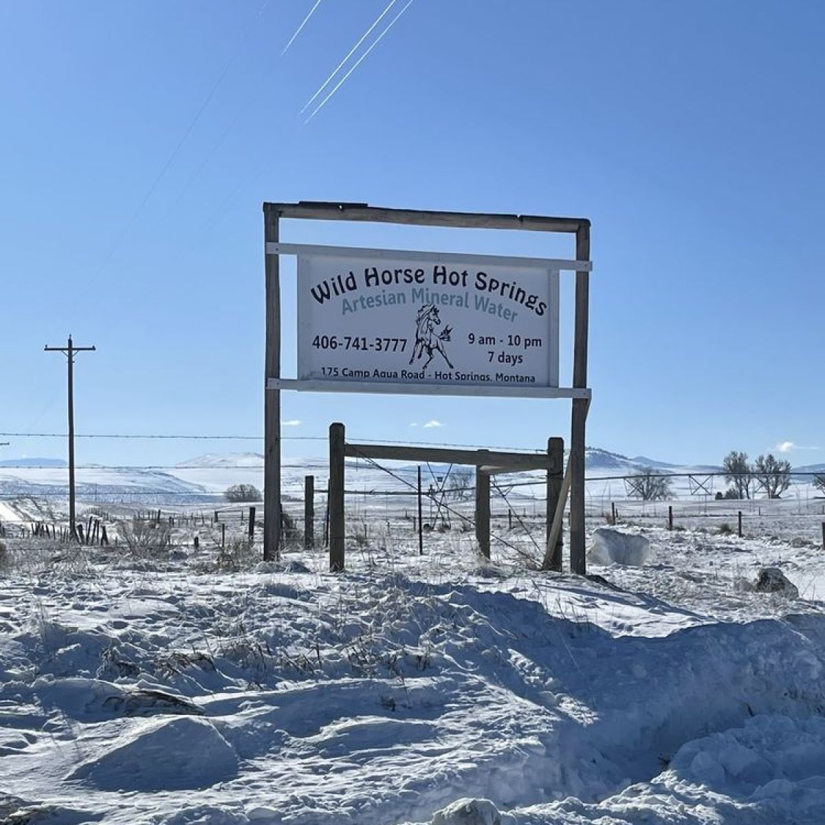 Snowy landscape with a sign for Wild Horse Hot Springs under a clear blue sky.